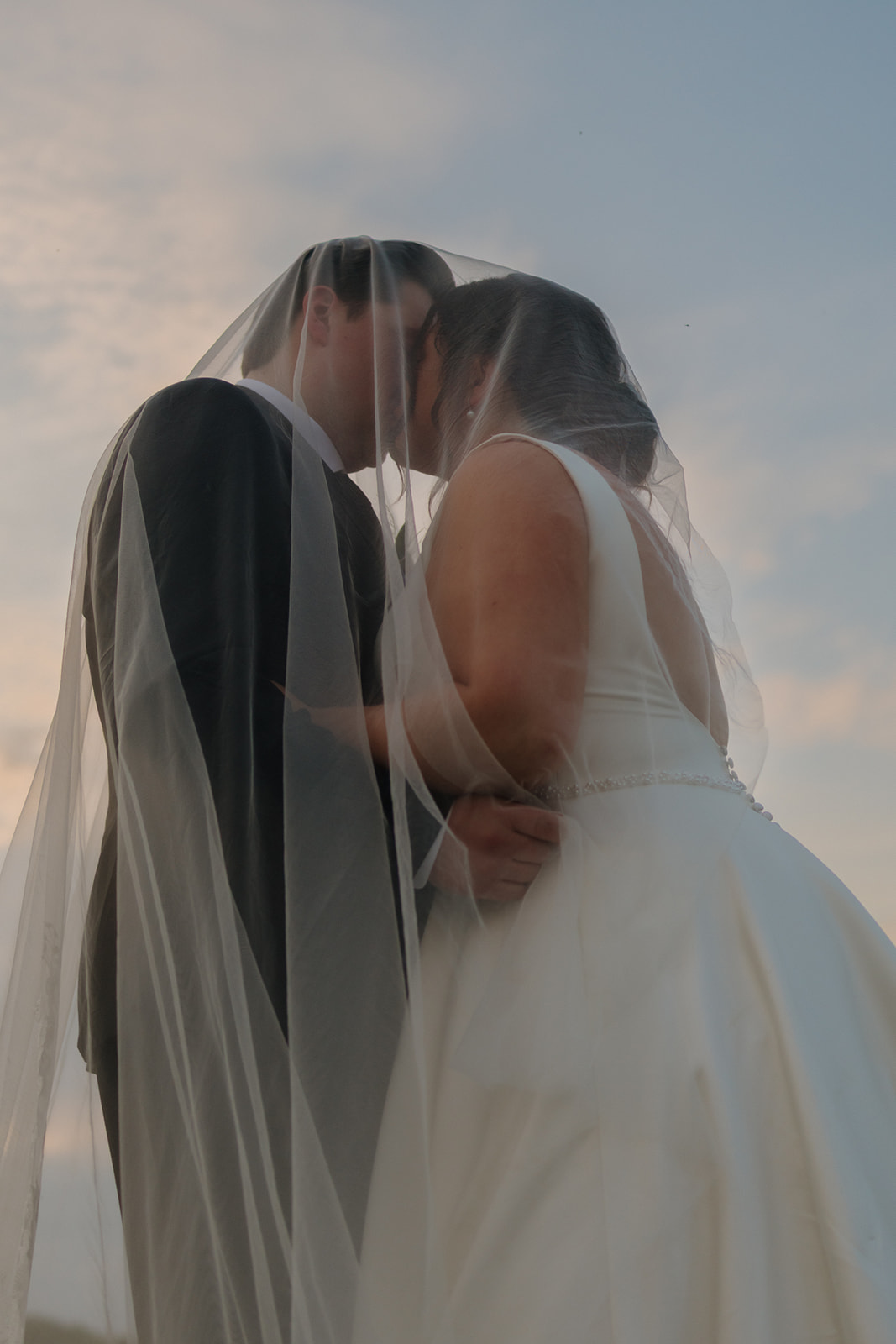 Romantic silhouette of a bride and groom kissing under her veil during golden hour, with soft skies behind them at their country club wedding.