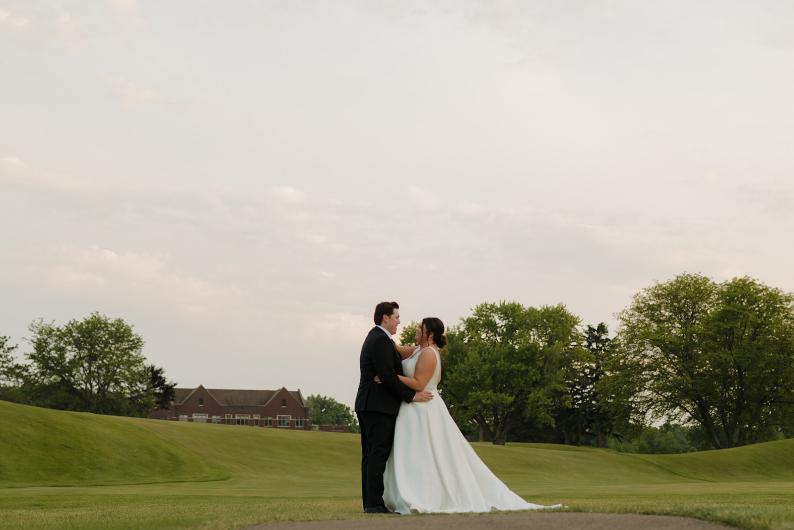 Bride and groom embracing on the green at sunset with the country club venue in the background, surrounded by soft rolling hills.