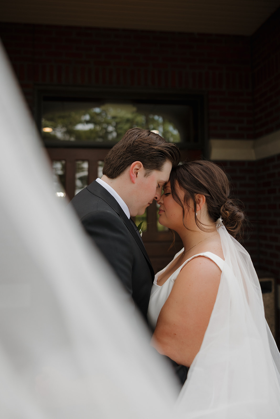 Intimate moment of a bride and groom standing close with foreheads touching, her veil softly framing them outside the country club entrance.