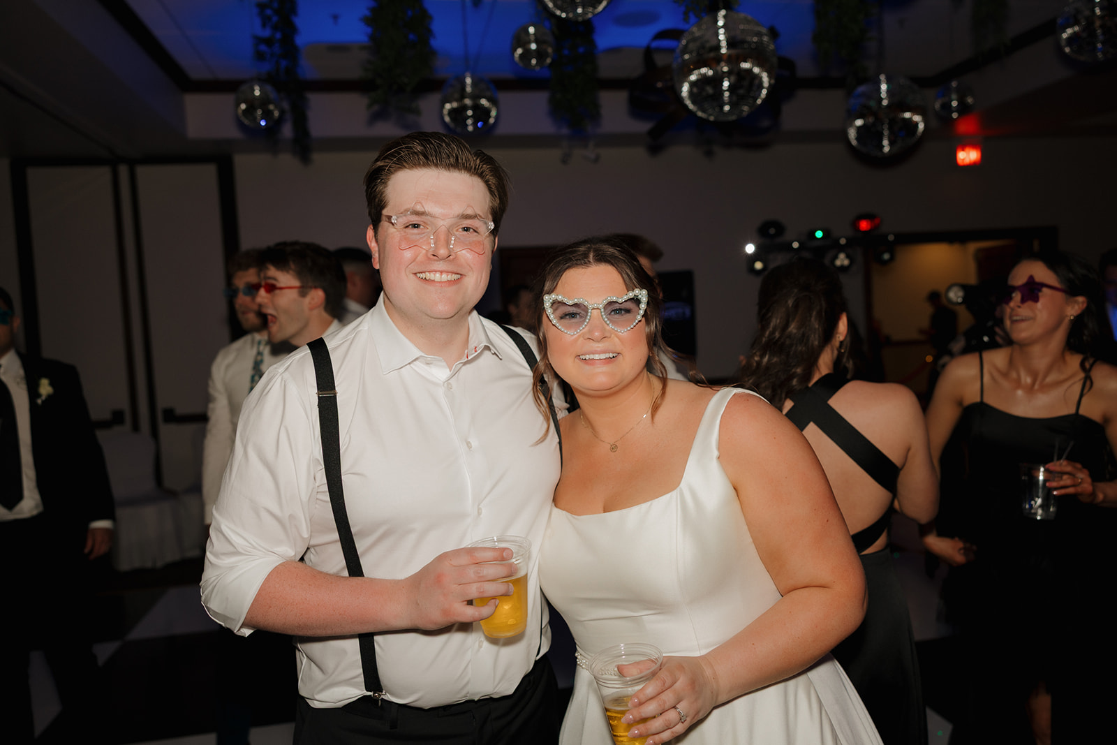 Bride and groom wearing fun party glasses and holding drinks while smiling on the dance floor during their country club wedding reception, with disco balls hanging overhead.