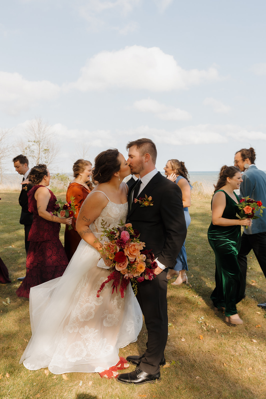 Bride and groom kiss as newly married couple surrounded by wedding party behind them