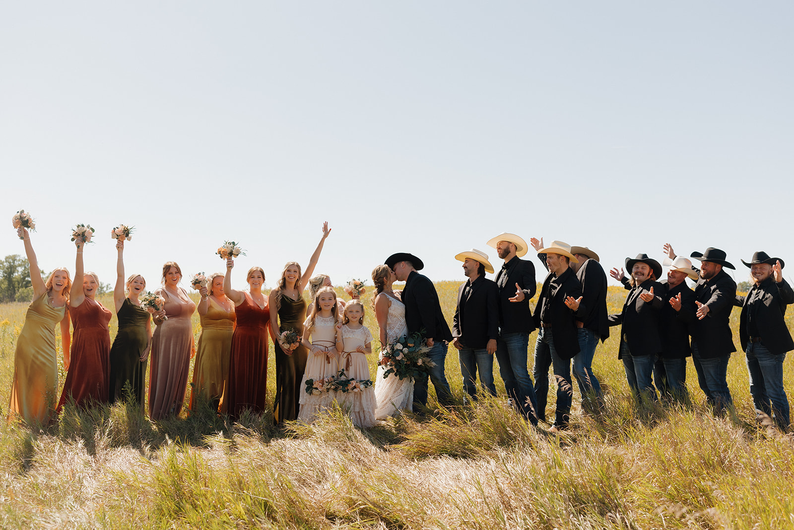 Wedding party cheers on the couple as they kiss in a golden field, with bridesmaids in warm-toned dresses and groomsmen rocking cowboy hats—pure backyard wedding joy.
