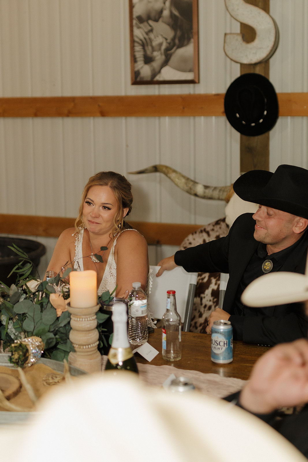 Bride gets emotional during speeches at the sweetheart table while the groom, in a cowboy hat, smiles beside her during their backyard wedding reception.