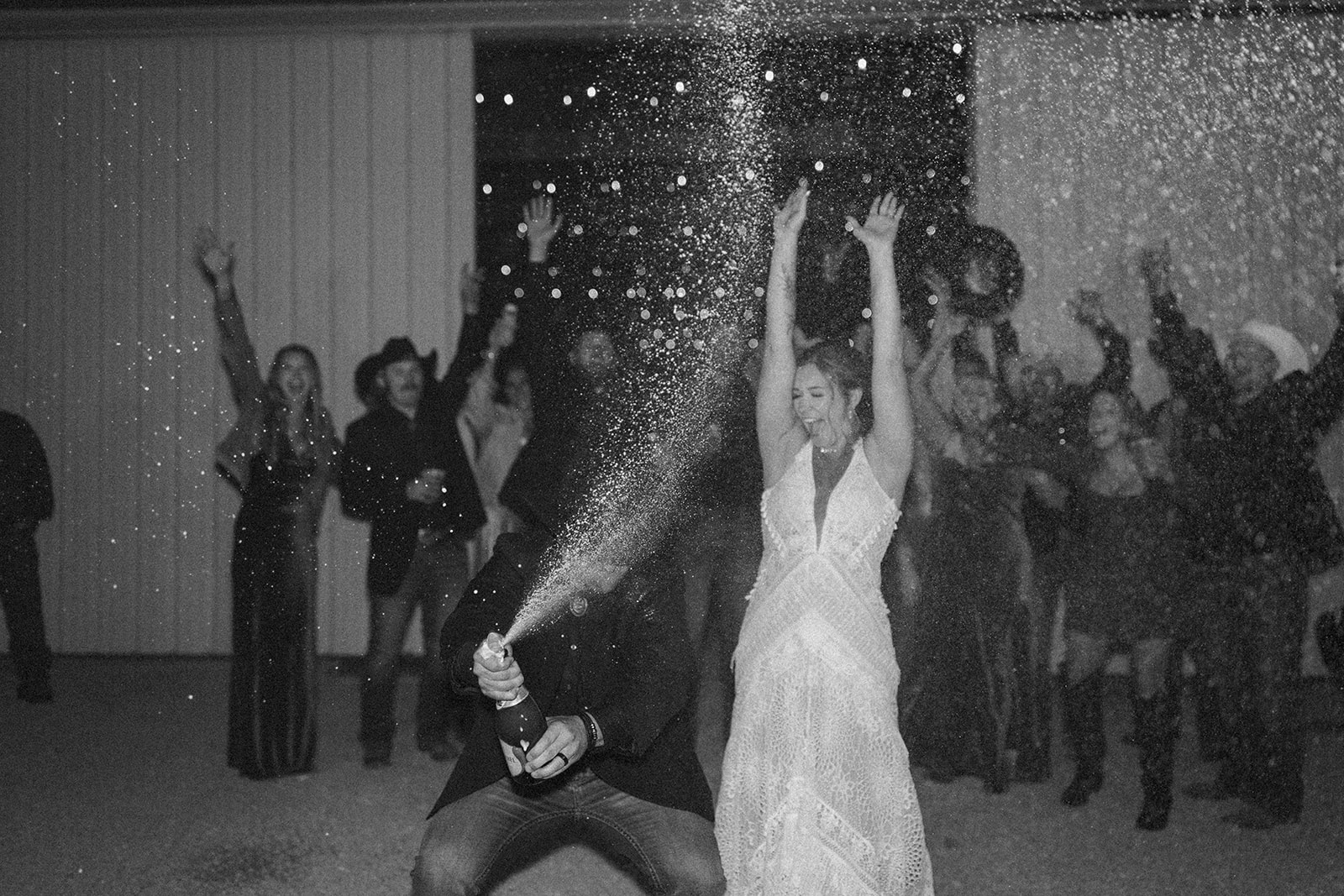 Black and white photo of the couple popping champagne outside their venue, surrounded by cheering guests under string lights.
