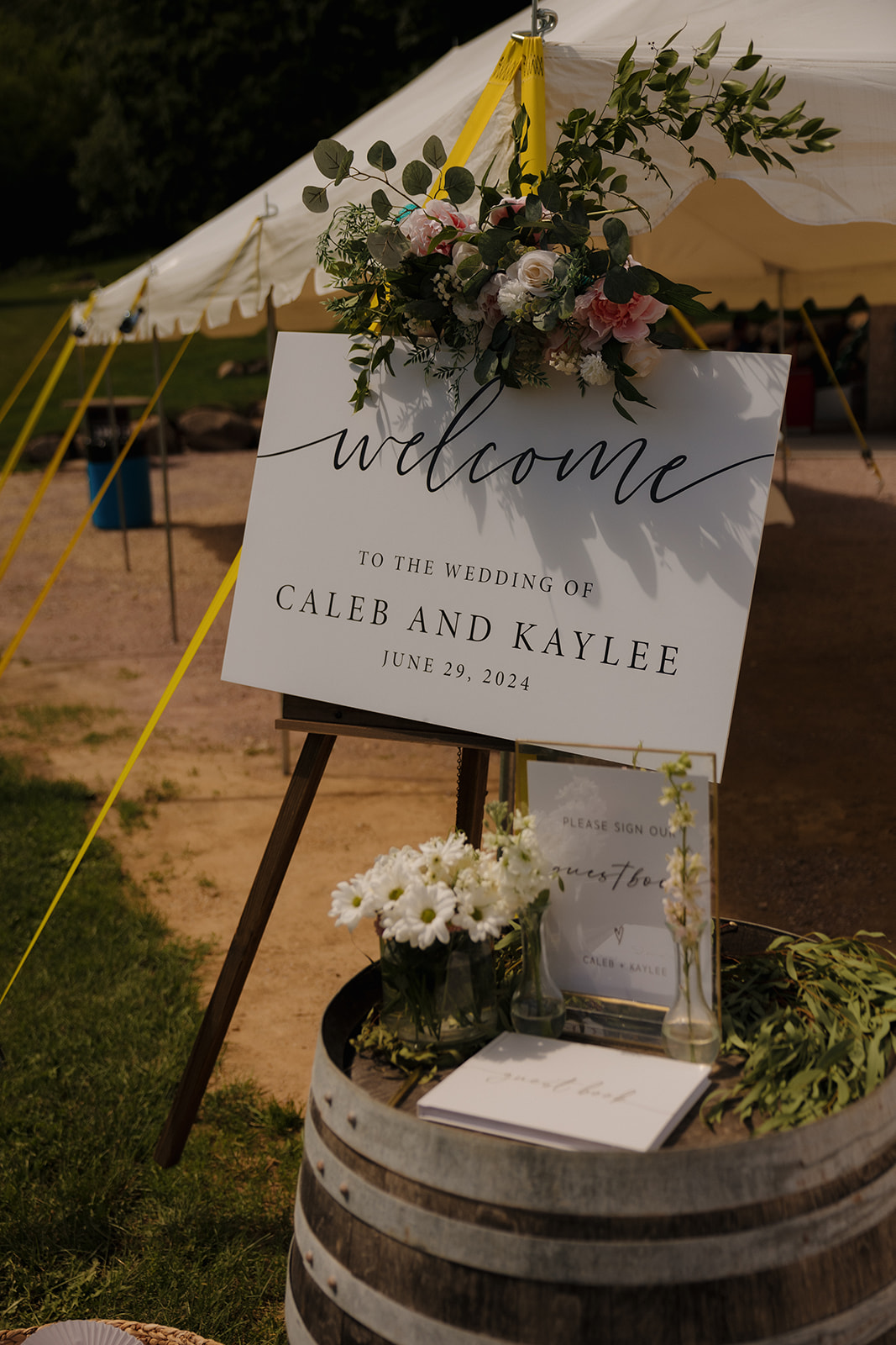 Elegant welcome sign for Caleb and Kaylee’s backyard wedding, displayed on a wine barrel with flowers and personal touches beneath a white tent.