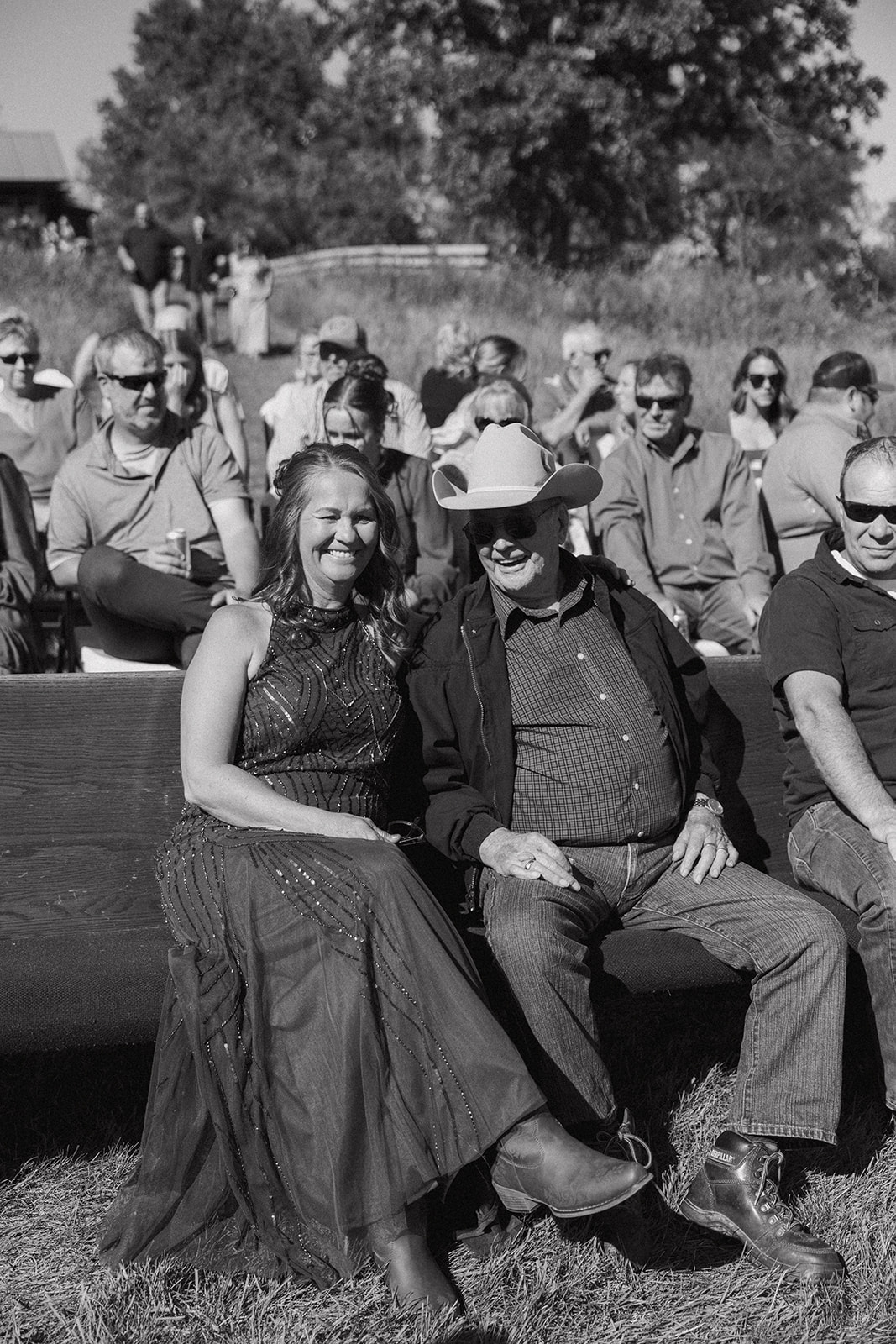 Black and white photo of smiling guests seated on wooden benches, enjoying the sunny outdoor ceremony at a country-inspired backyard wedding.