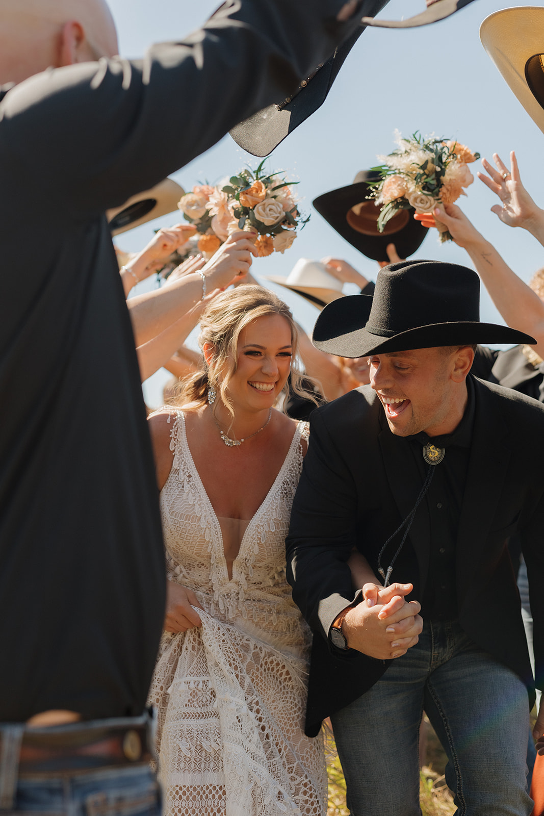 Bride and groom laugh as they run through a cowboy hat and bouquet tunnel made by their wedding party during a fun backyard wedding celebration.