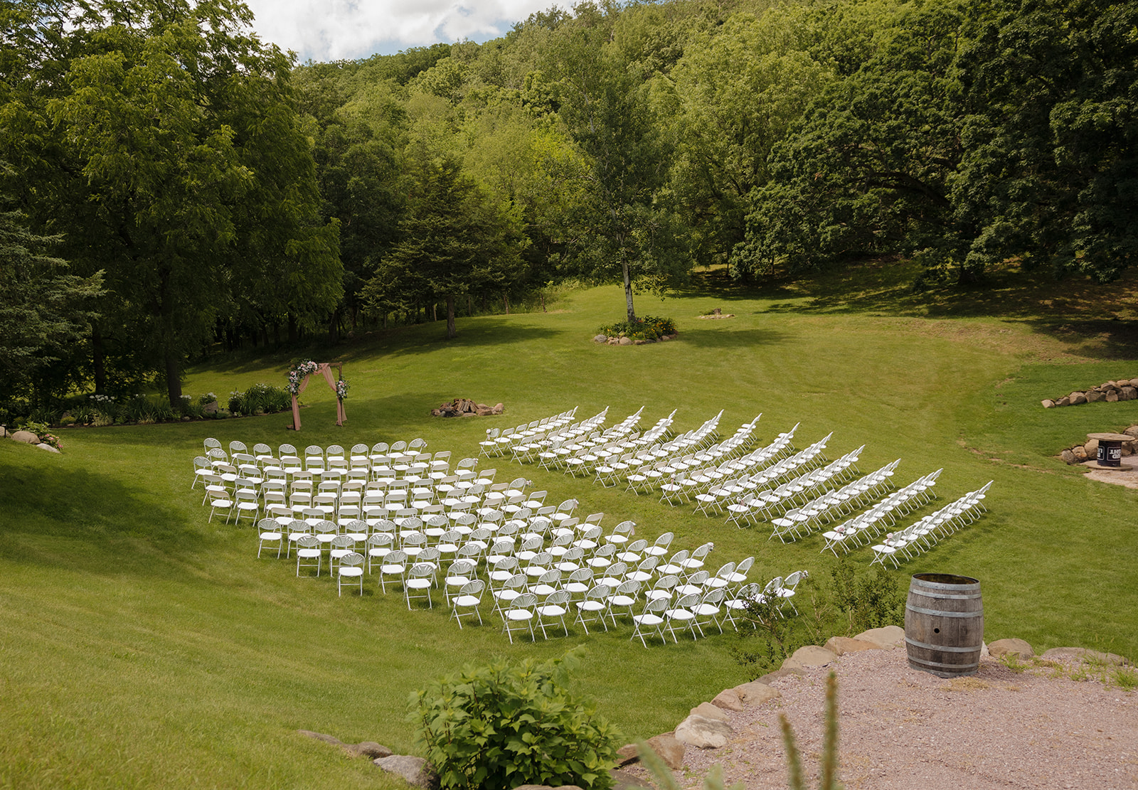 Wide view of a lush green backyard wedding ceremony setup with curved rows of white chairs and a floral arch tucked into a forested clearing.