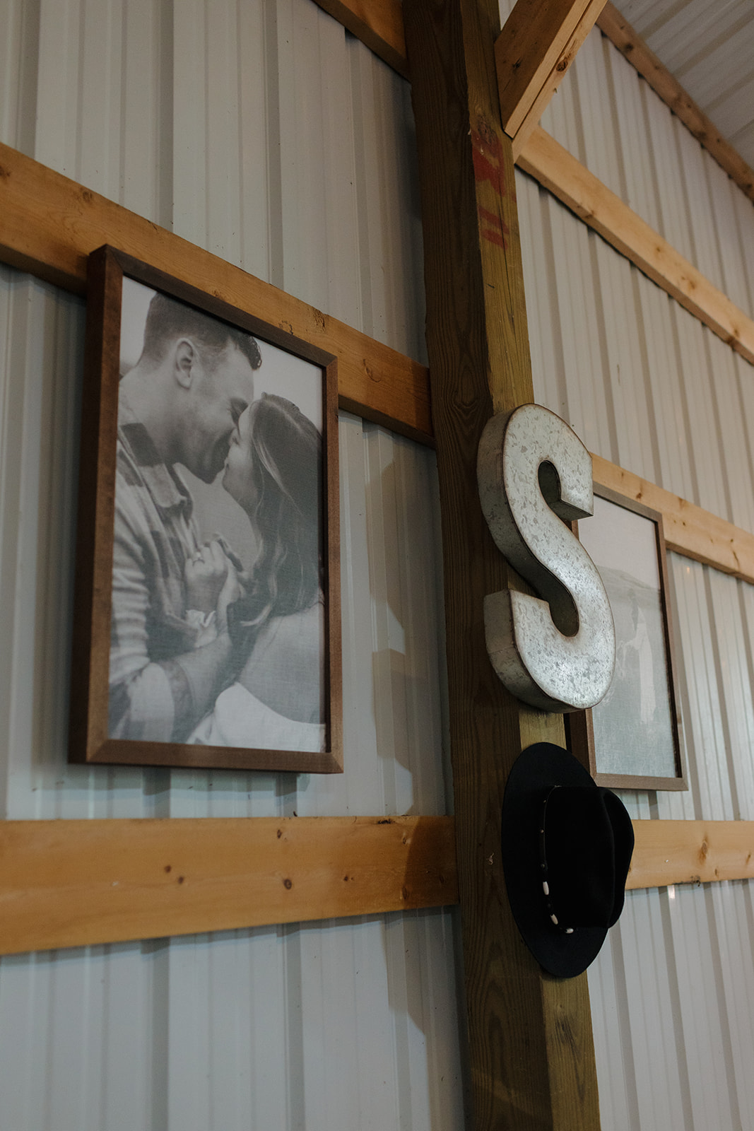 Rustic wall decor featuring engagement photos, a large galvanized “S,” and a cowboy hat hanging inside a cozy barn reception space.