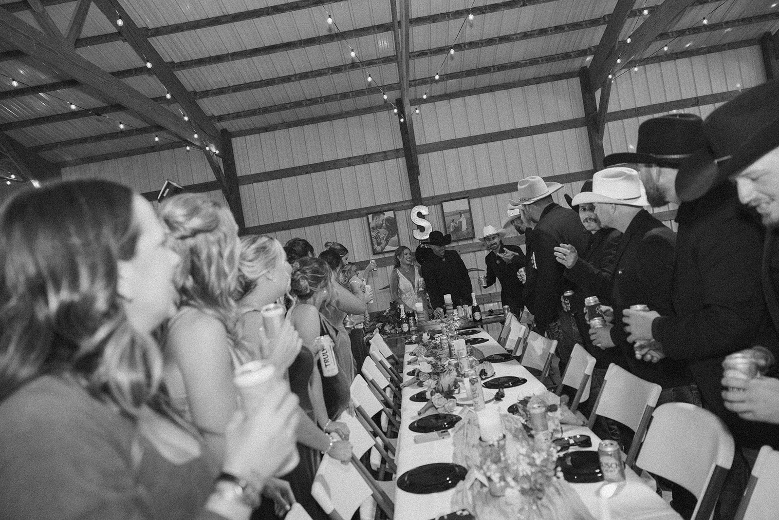 Black and white image of the wedding party cheering and raising drinks as the newlyweds enter the barn-style reception at their backyard wedding.