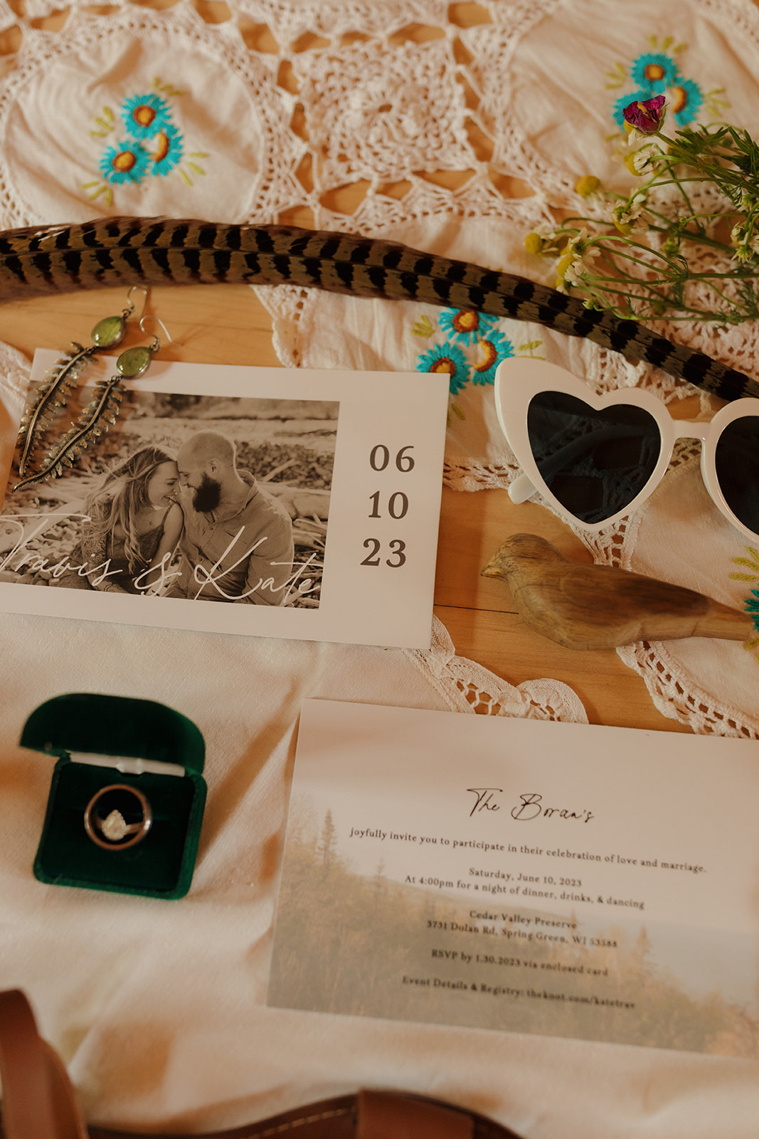 A flat lay of wedding details including heart-shaped sunglasses, a velvet ring box, feathers, and a photo save-the-date on a vintage tablecloth.