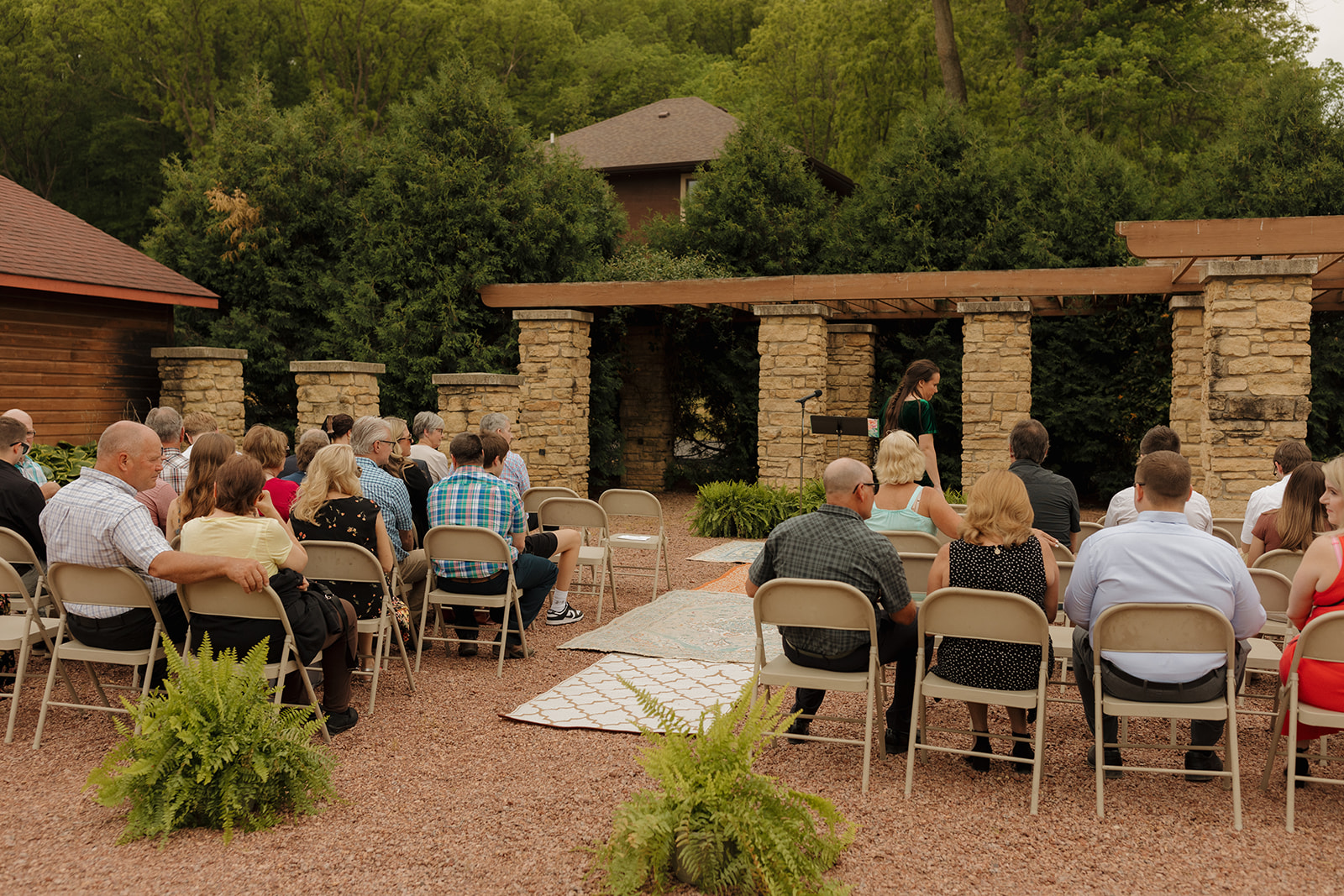 Guests begin to take their seats at a rustic outdoor venue surrounded by trees and stone columns, waiting for the backyard wedding ceremony to begin.