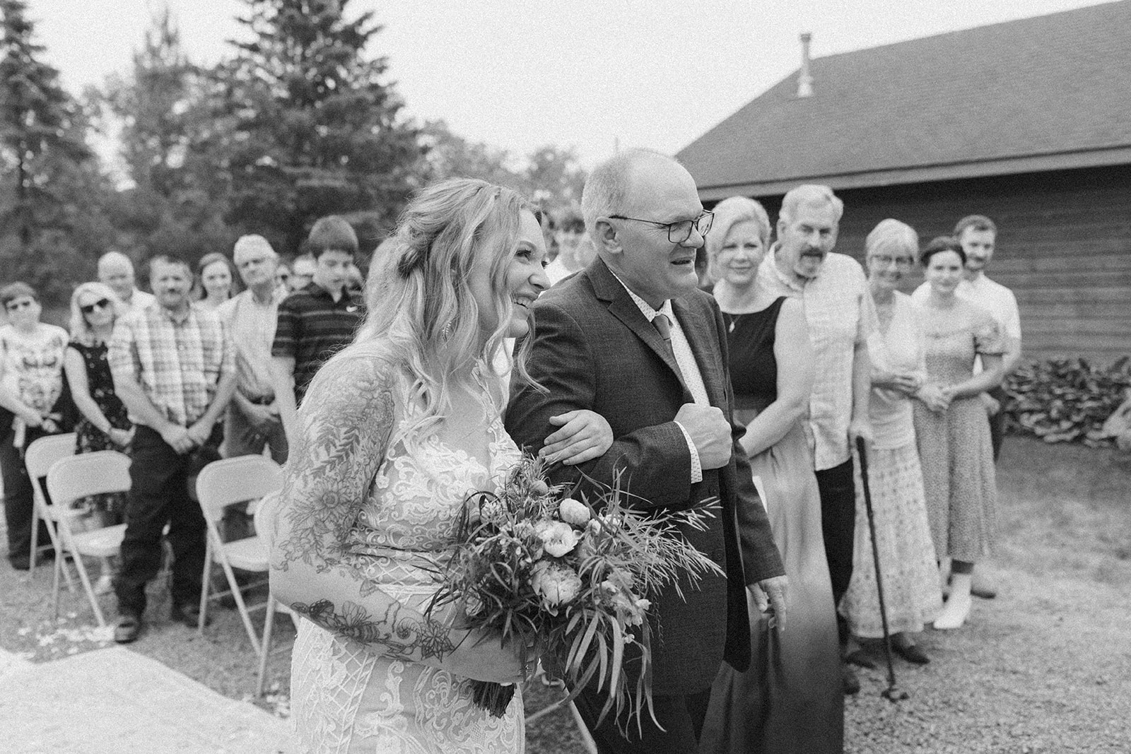 Bride and her father walk down the aisle arm in arm, surrounded by loved ones smiling and watching the ceremony unfold.