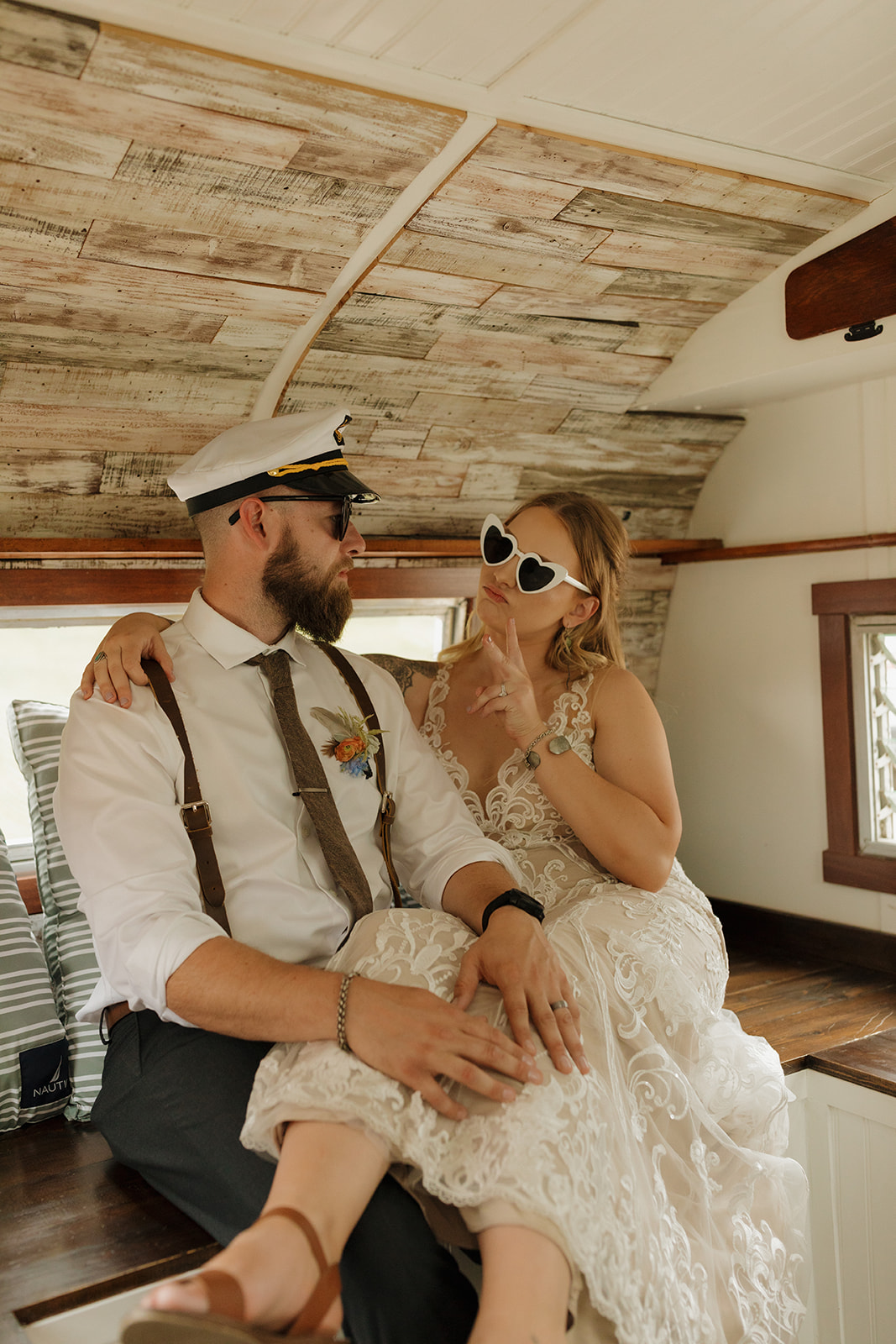 Bride and groom kick back inside a vintage camper, wearing heart sunglasses and a captain's hat, sharing a quiet moment of fun during their big day.