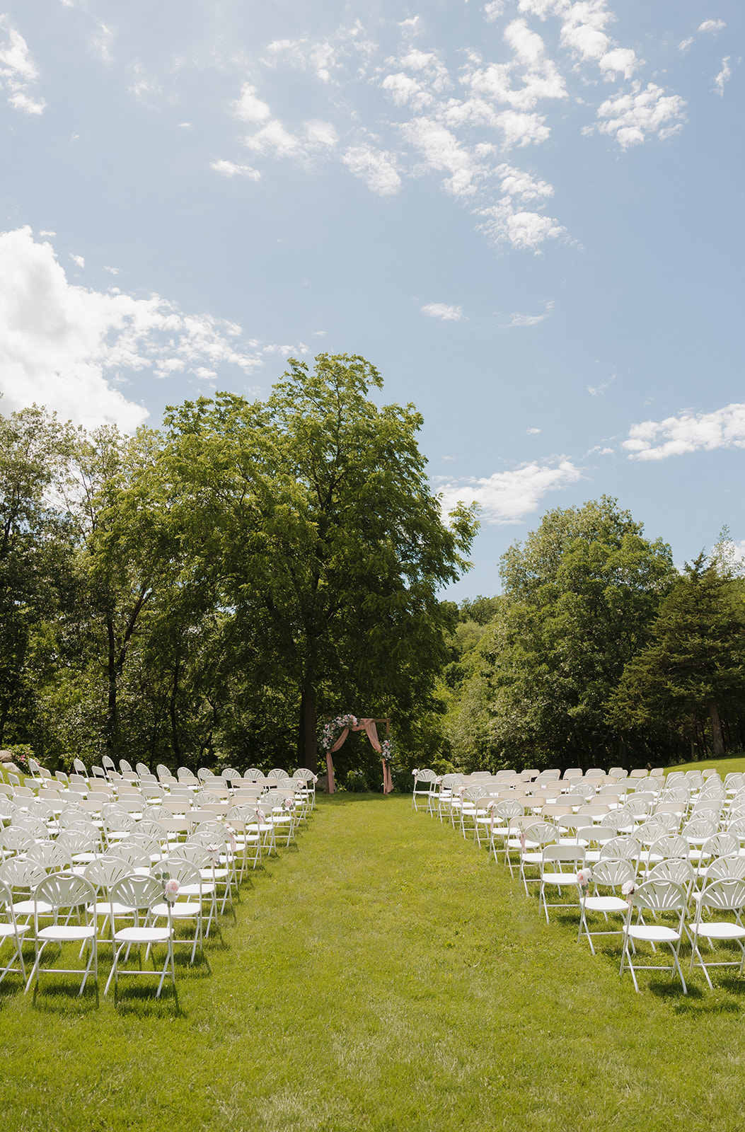 A peaceful ceremony setup with rows of white chairs facing a floral arch under tall trees on a bright, sunny day.