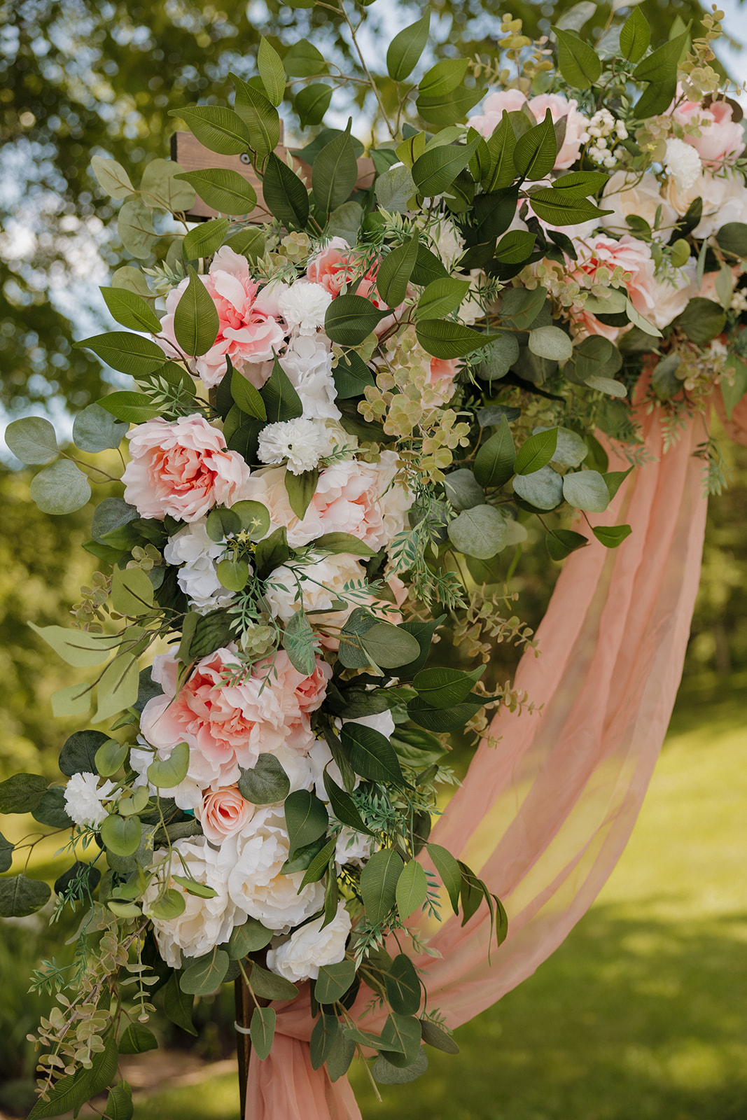 Close-up of a floral ceremony arch featuring lush greenery and soft blush, white, and peach flowers with flowing pink chiffon.