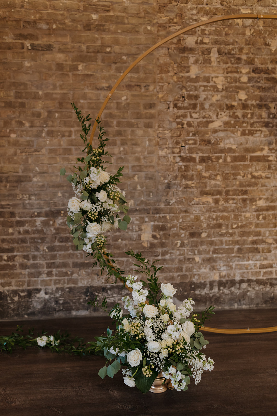 Curved floral arch with white roses and greenery set against a rustic brick wall at The Gage.