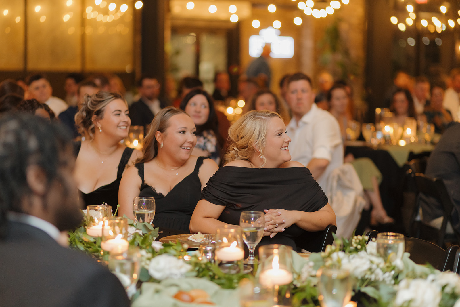 Wedding guests smiling and enjoying heartfelt reception speeches under twinkle lights.