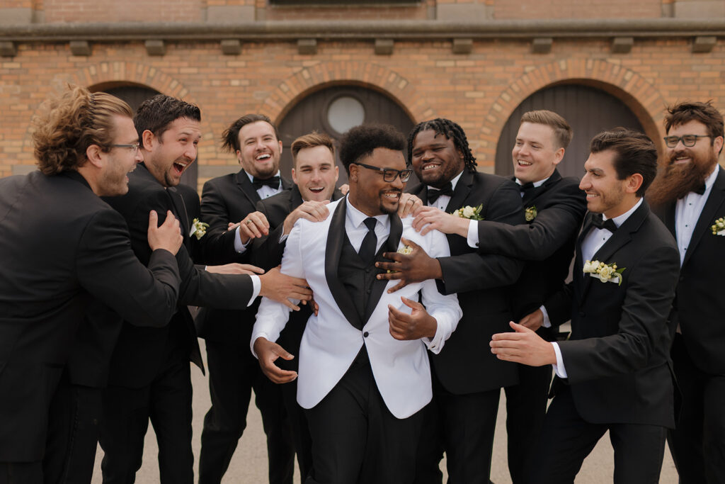 Groom surrounded by groomsmen in tuxedos, laughing during portraits before the wedding.