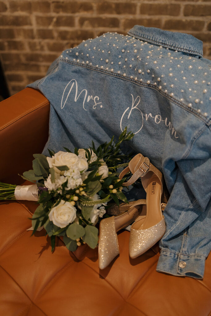 Personalized "Mrs. Brown" denim jacket paired with bouquet and bridal heels on a leather chair.
