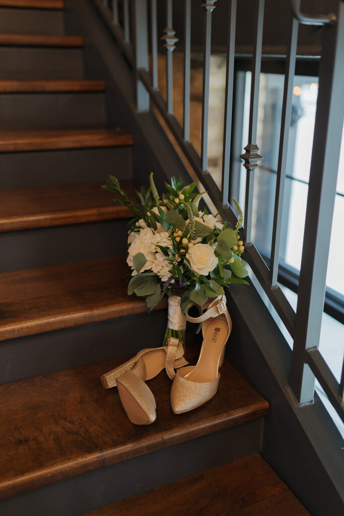 Gold bridal heels and white rose bouquet resting on wooden stairs at The Gage wedding venue.