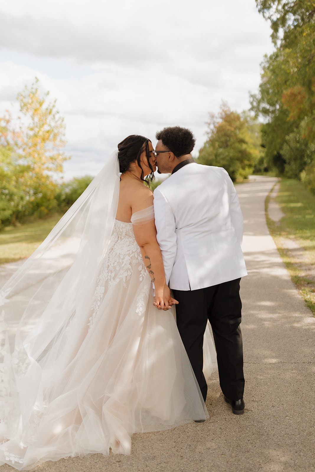 Bride and groom share a kiss while walking a tree-lined path near Lake Michigan.