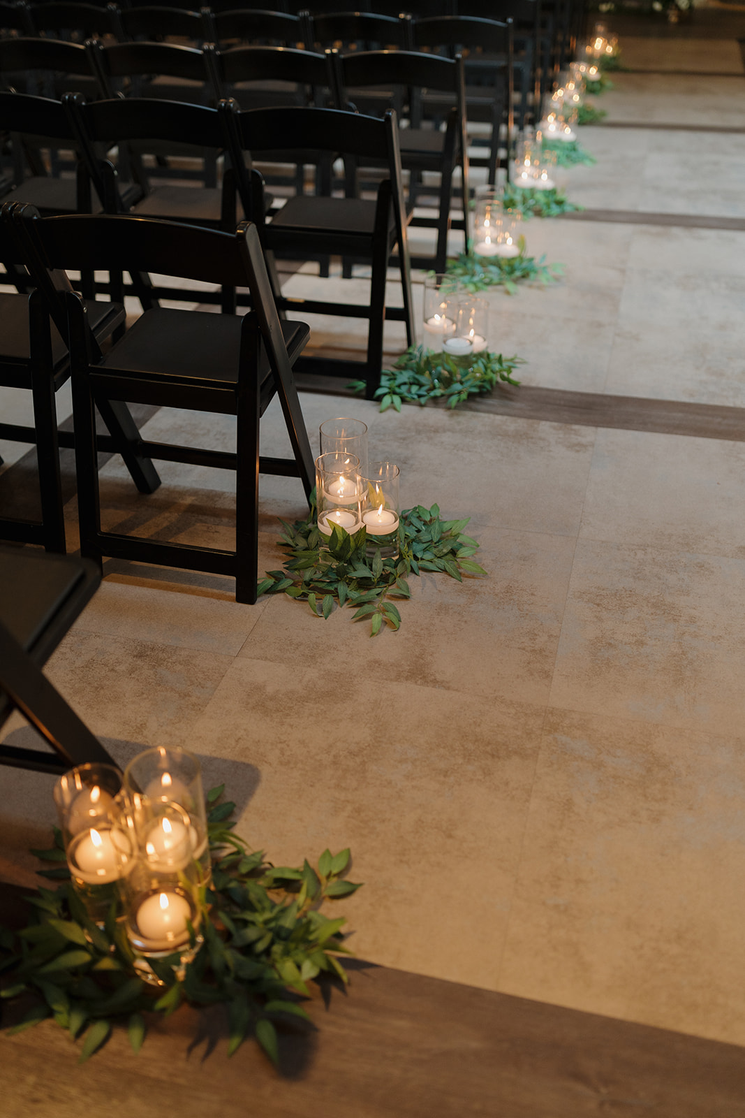 Glass candle holders and greenery line the ceremony aisle for a romantic indoor wedding setup.