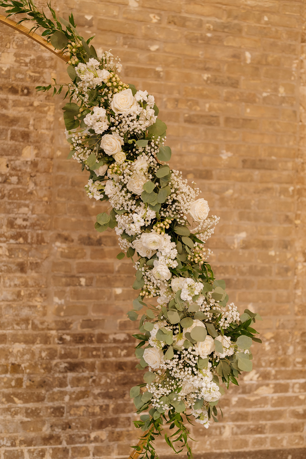 Asymmetrical floral arch with white roses and eucalyptus, styled against an industrial brick wall.