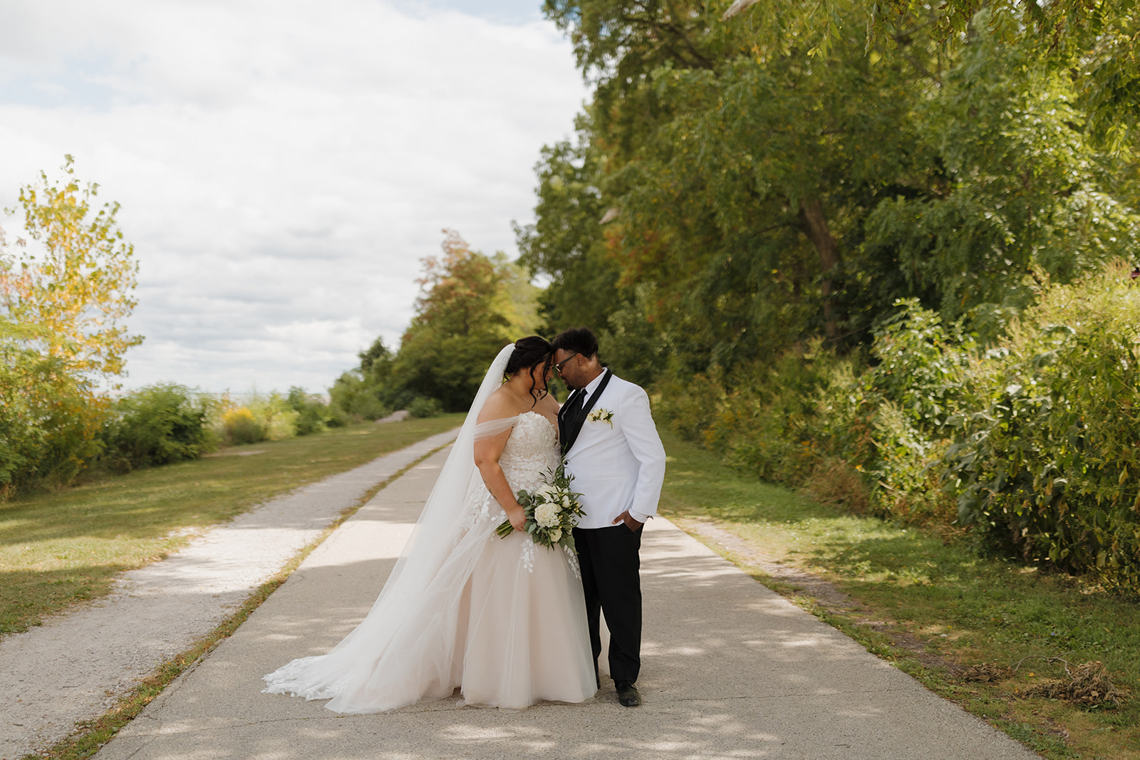 Bride and groom walking along a tree-lined lakefront path during their wedding portraits in Milwaukee.