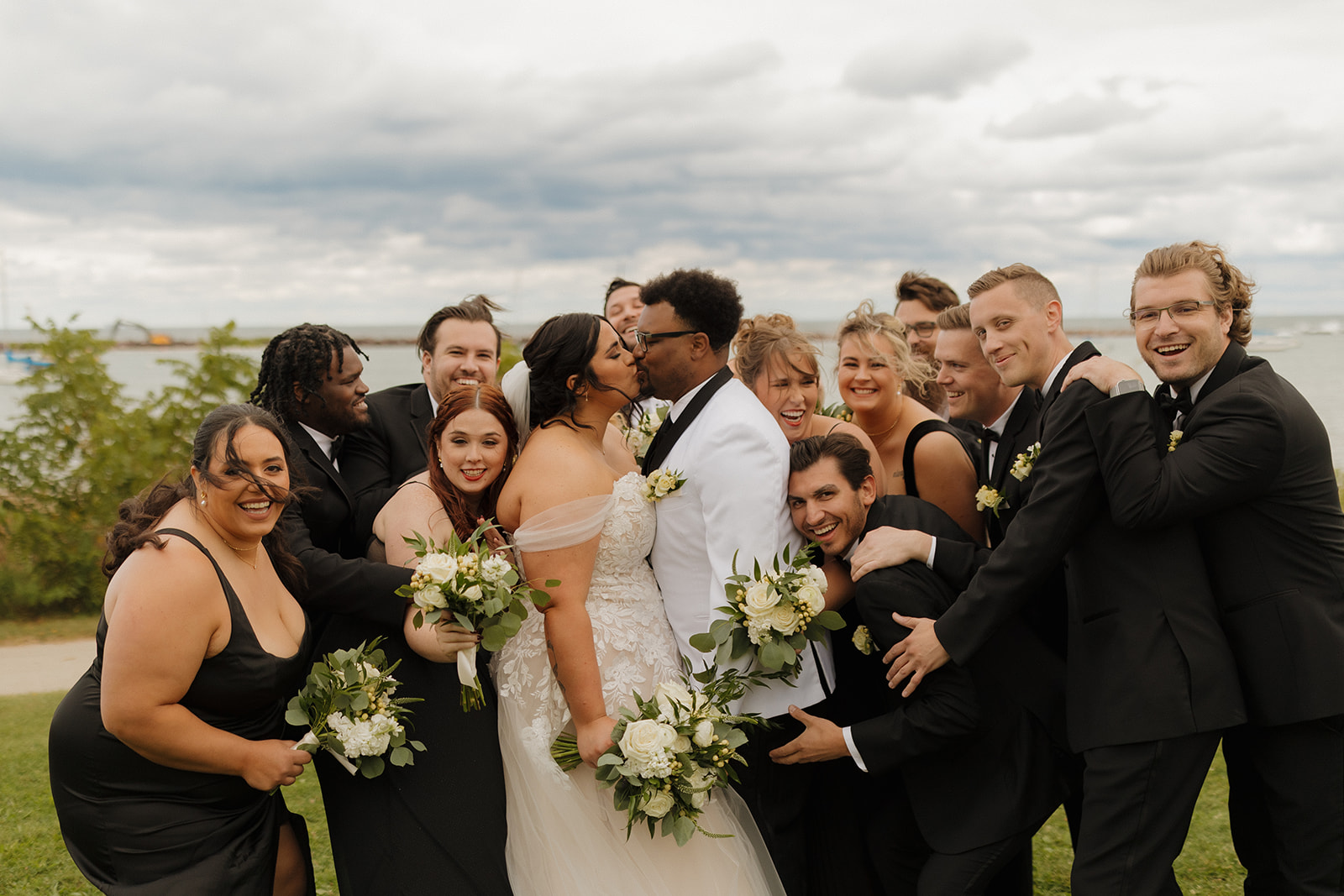 Wedding party laughing and hugging the couple as they kiss during portraits by Lake Michigan.