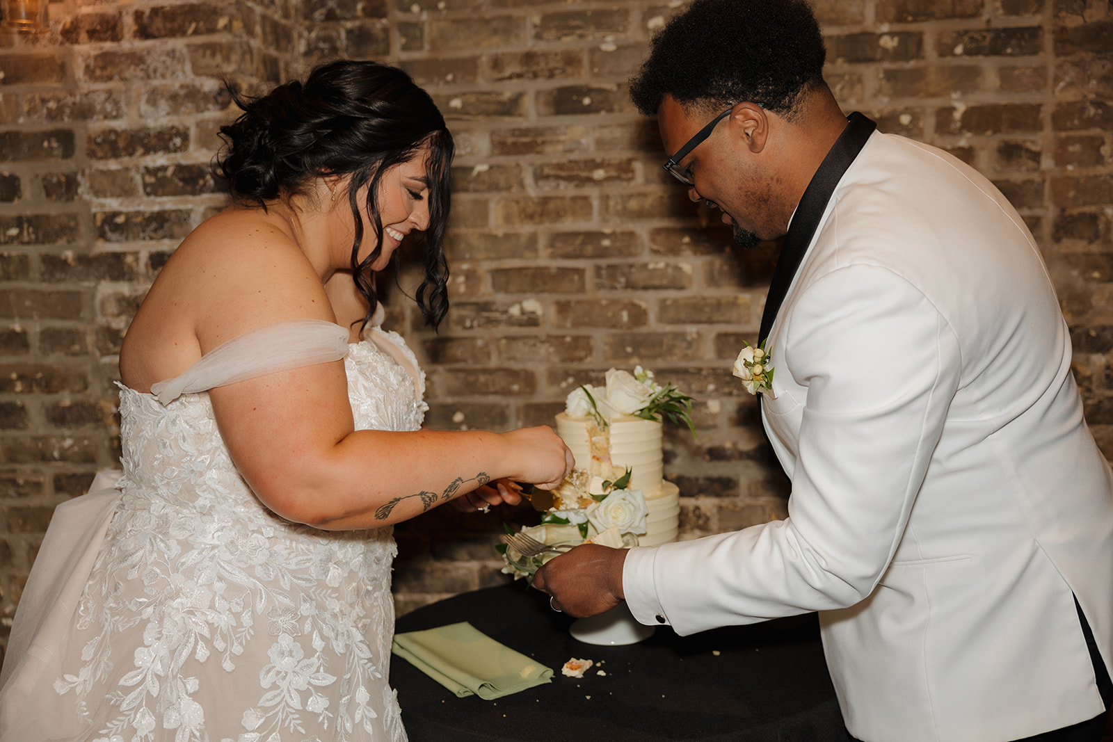 Bride and groom laughing as they cut their white wedding cake decorated with roses and greenery.