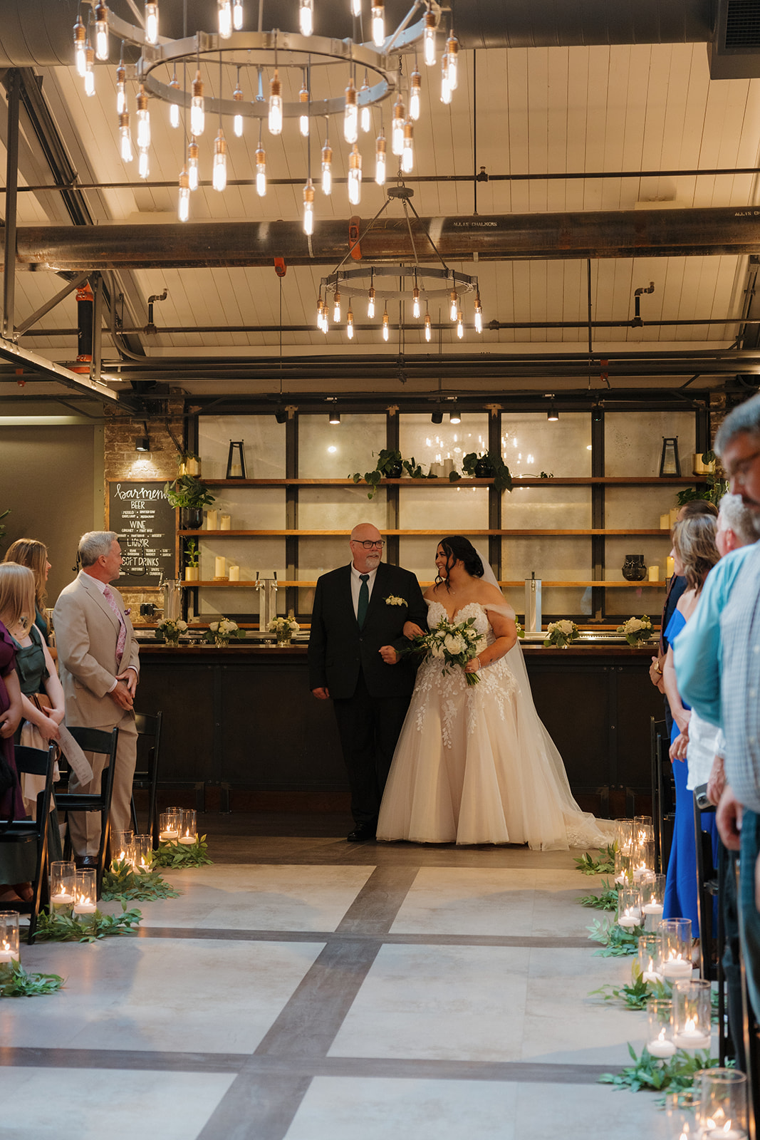 Bride walks down a candlelit aisle with her father during an indoor ceremony.