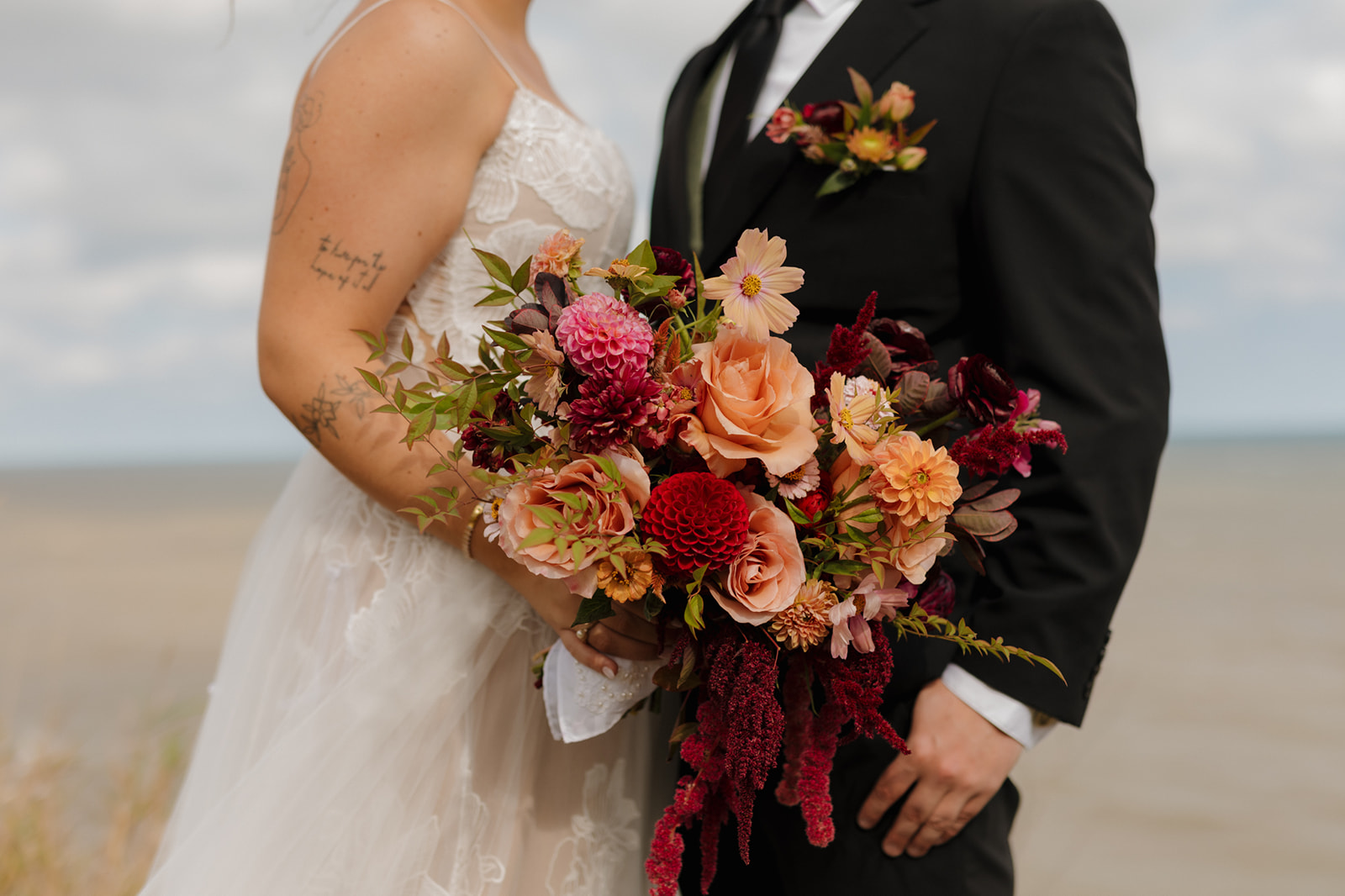Close-up of bride’s textured lace gown and bold fall bouquet next to groom’s green vest, with lake views at their Wisconsin wedding.