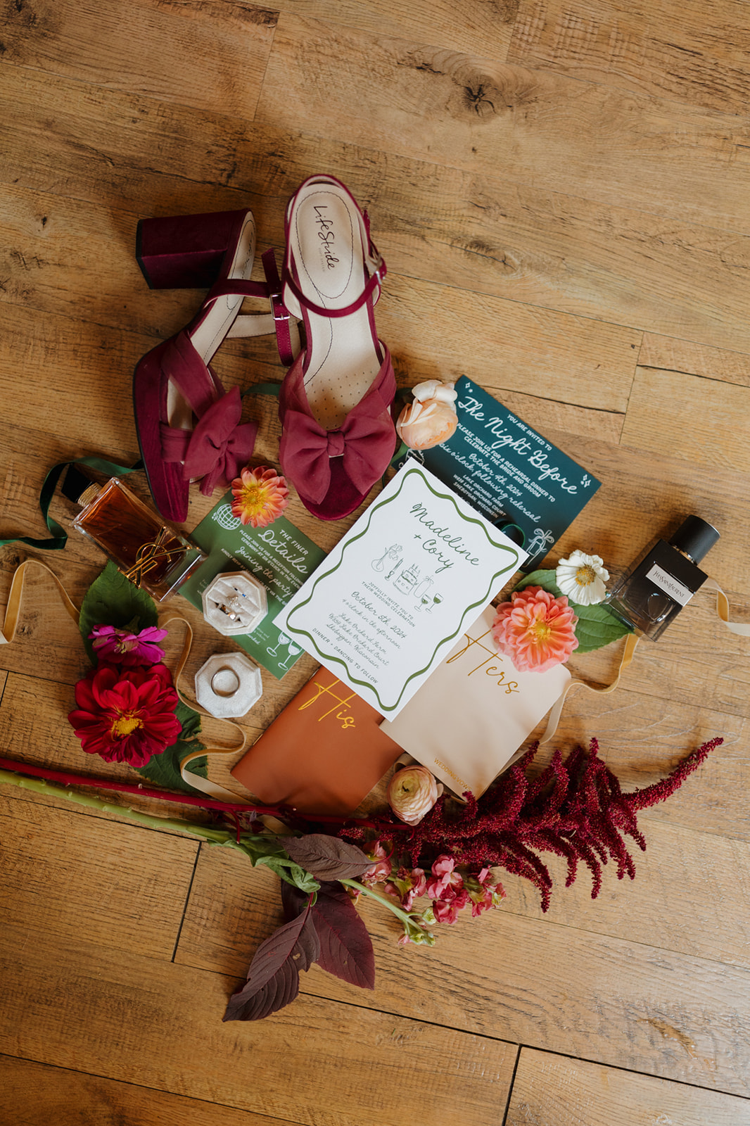 Flatlay of Wisconsin wedding details including burgundy bridal heels, vow books, floral accents, invitations, and signature scent on wood flooring.