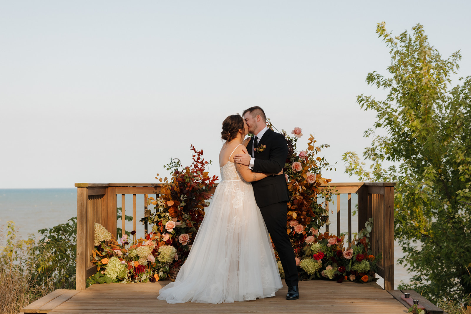 Bride and groom share a kiss at their lakeside ceremony, surrounded by fall-toned floral arrangements during their Wisconsin wedding.