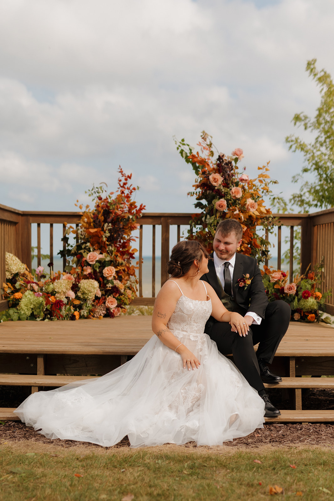 Newlyweds sit hand-in-hand on wooden altar steps, surrounded by bold florals and lake views at their colorful Wisconsin wedding.