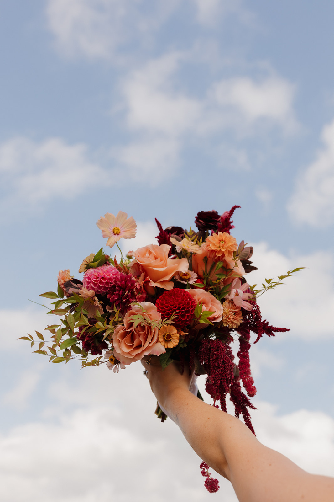 Vibrant bridal bouquet held up against a blue sky, featuring rich pink, peach, and burgundy florals for a colorful Wisconsin wedding.