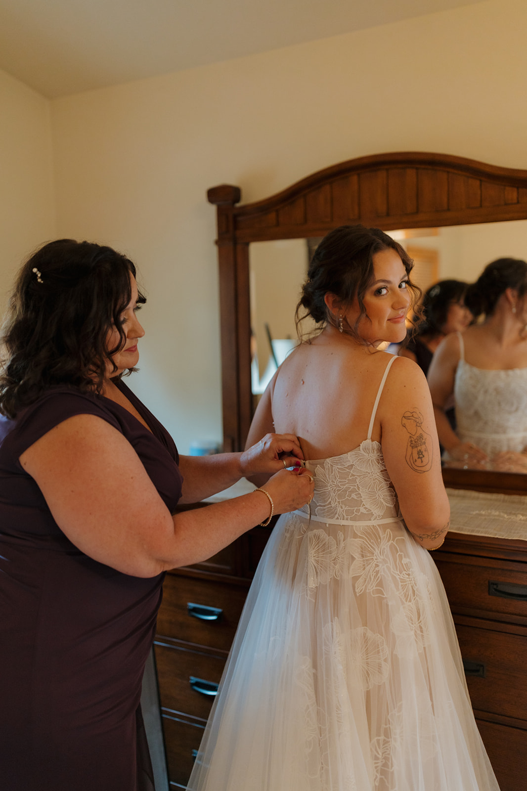 Bride smiles over her shoulder while her mom helps with her dress in a cozy getting-ready room on the morning of a Wisconsin wedding.
