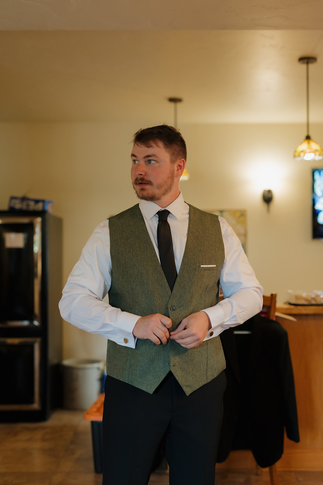 Groom buttons his green vest while getting ready indoors, preparing for his Wisconsin wedding with calm anticipation.