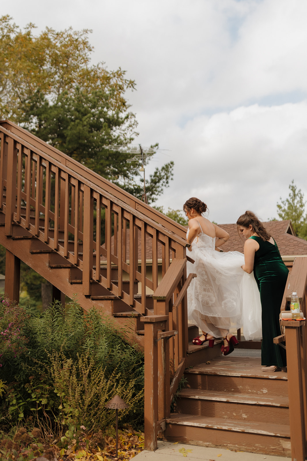Bride walks up a wooden staircase with the help of her bridesmaid, showing off her lace gown and red heels before a lakeside Wisconsin wedding.