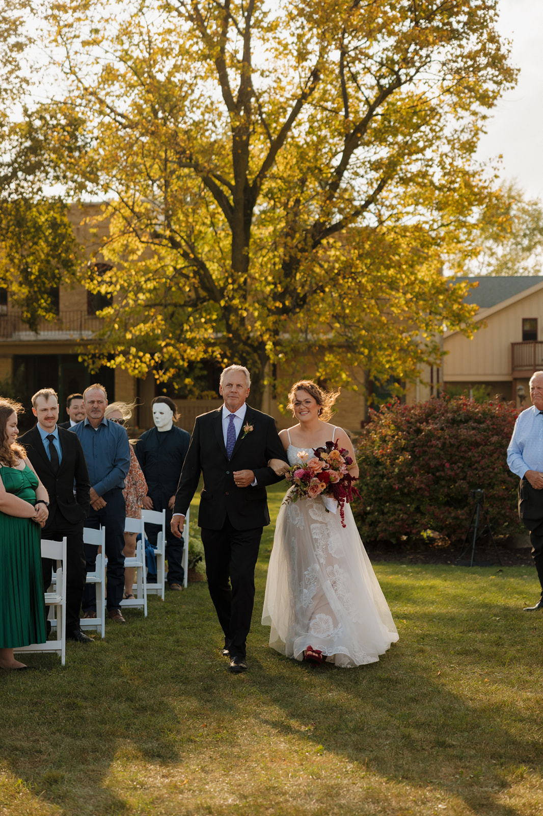 Bride walks arm-in-arm with her father down a grassy aisle lined with white chairs and golden autumn trees at a Wisconsin wedding ceremony.