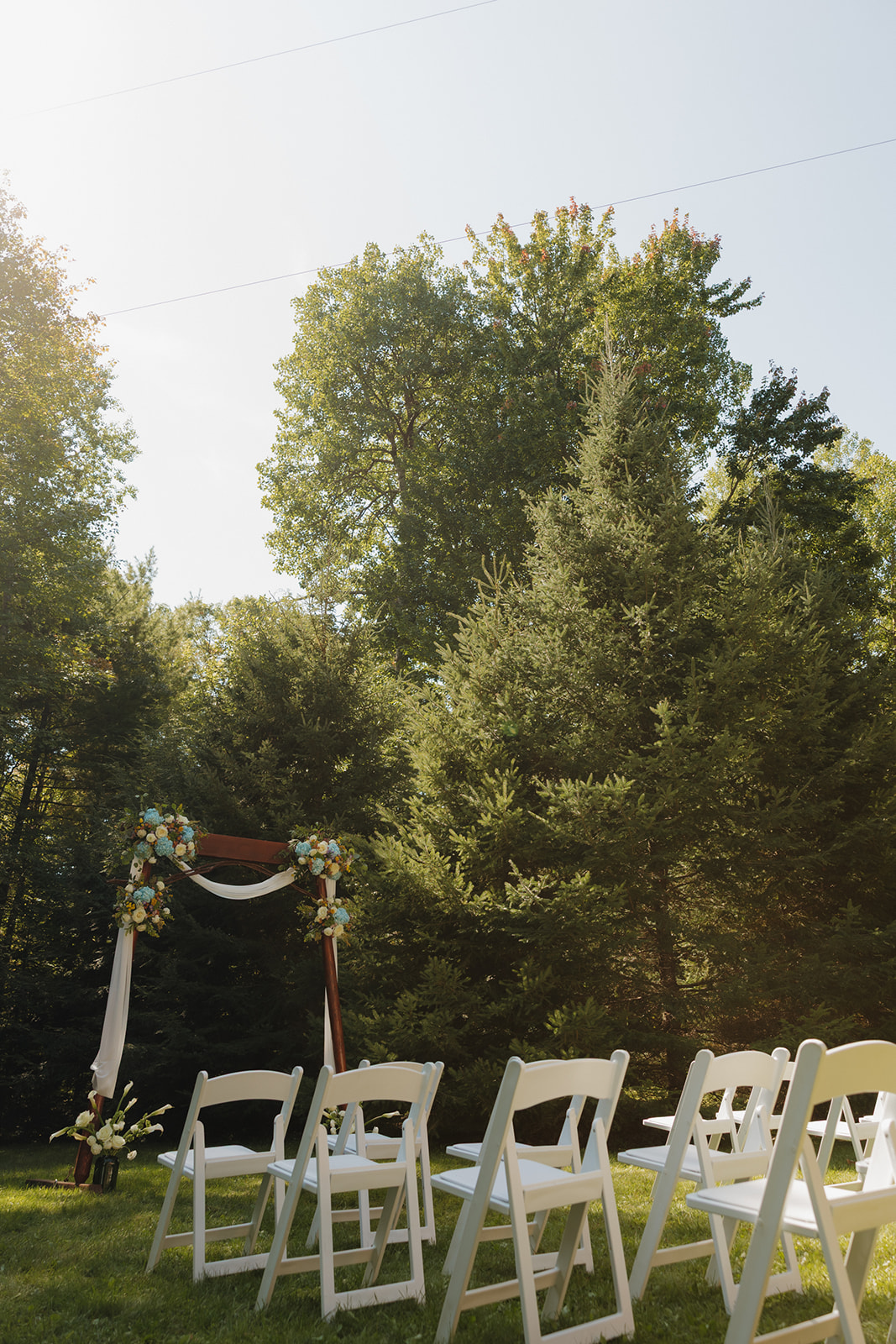 Rows of white chairs set up in a forest clearing with a floral arch for a barn wedding ceremony.