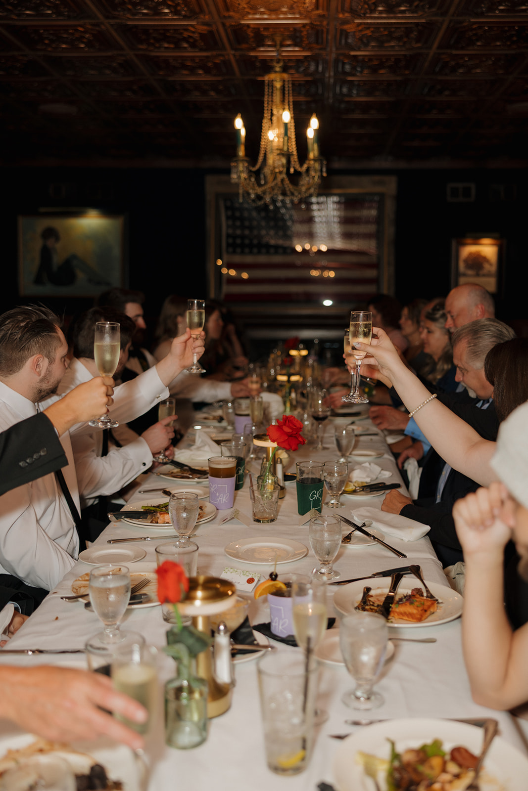 Guests raise champagne glasses for a toast at a long reception table with chandeliers overhead.