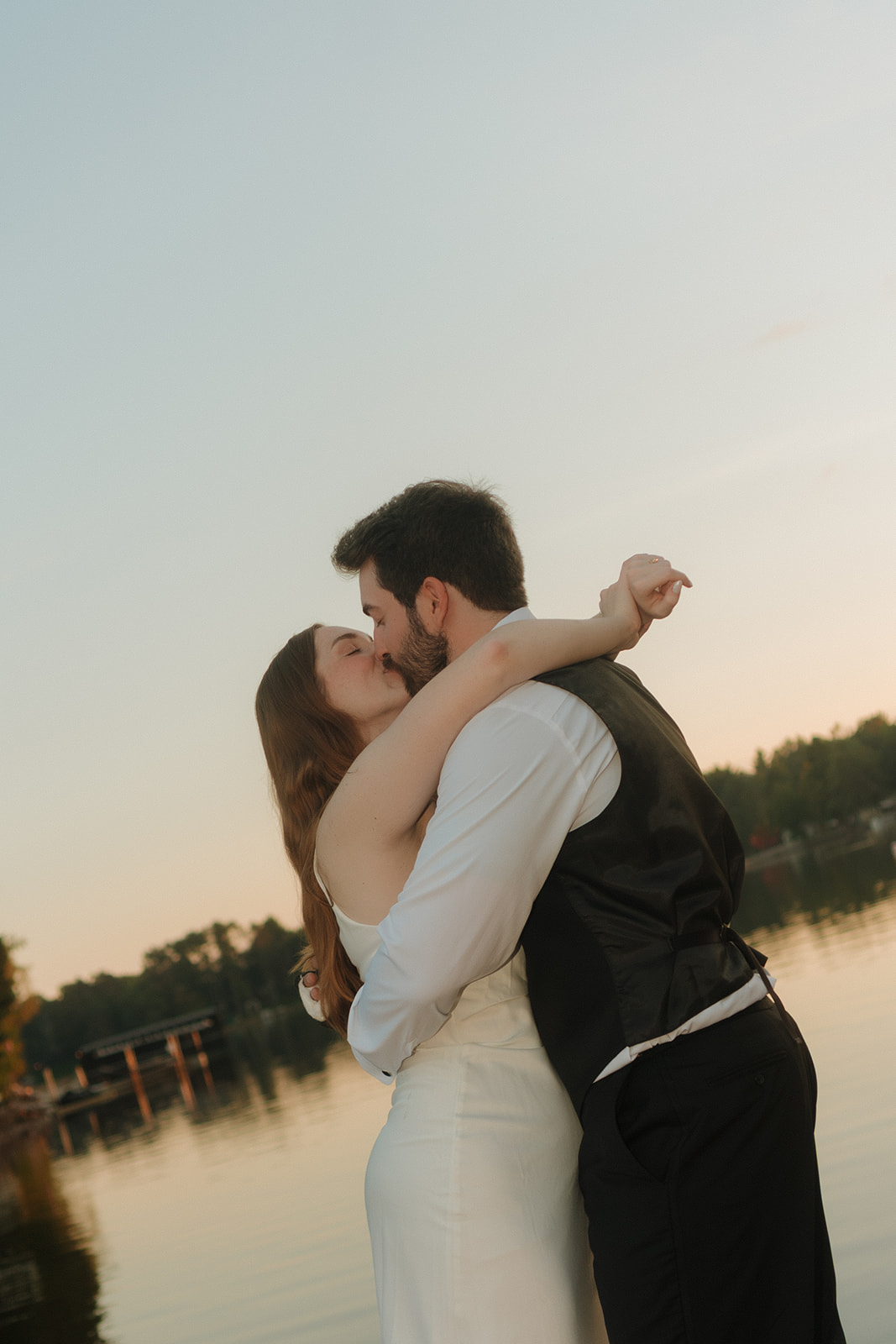 Bride and groom sharing a kiss at sunset by the lake, their reflection glowing on the water.