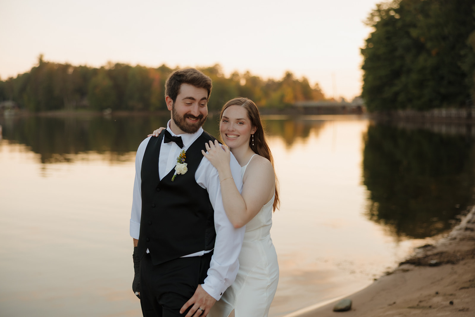 Bride and groom smiling together by the water at sunset during their barn wedding celebration.