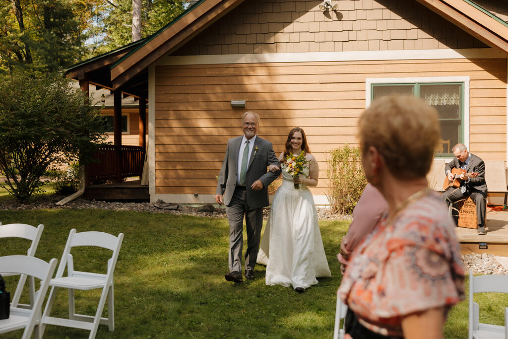 Bride walking down the aisle with her father, passing rustic cabins and guests at an outdoor ceremony.