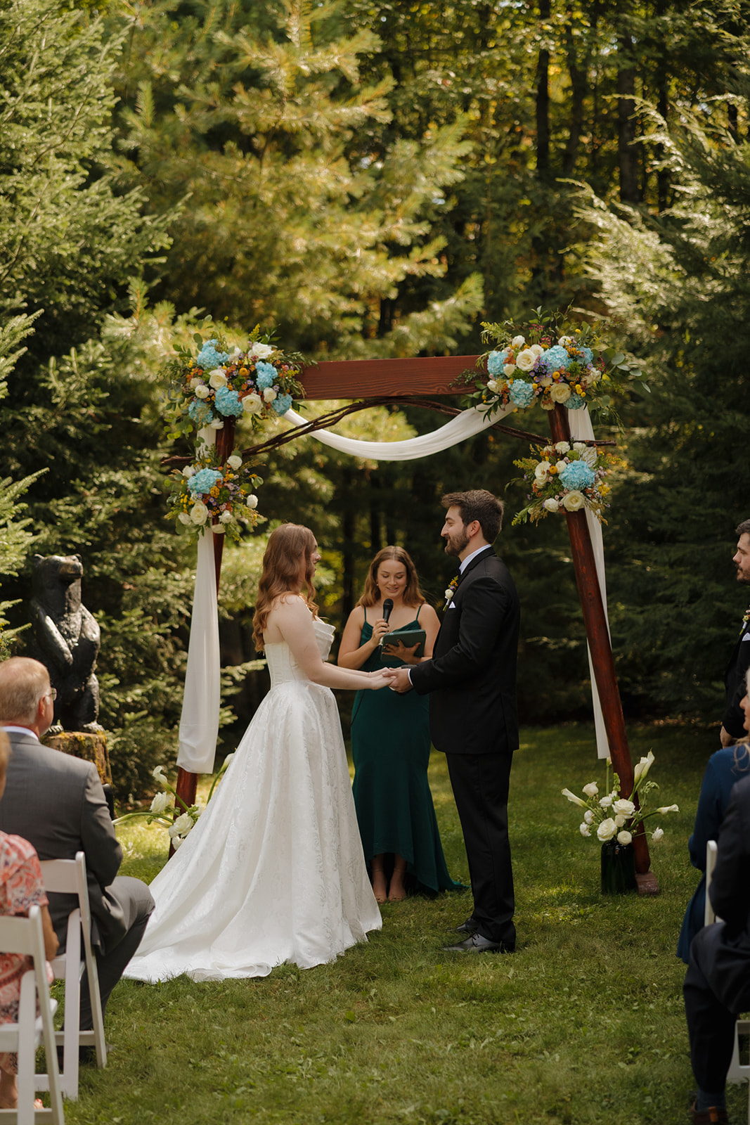 Bride and groom holding hands beneath a decorated arch while the officiant speaks.
