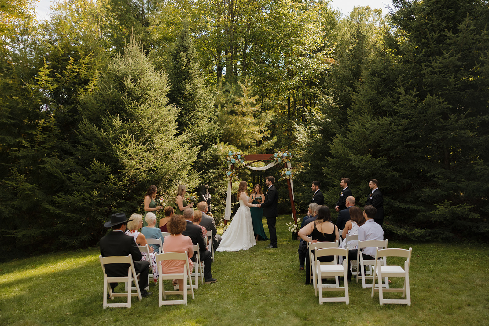 Wide view of a wedding ceremony in the forest, with guests seated in rows and the couple standing beneath a wooden arch.