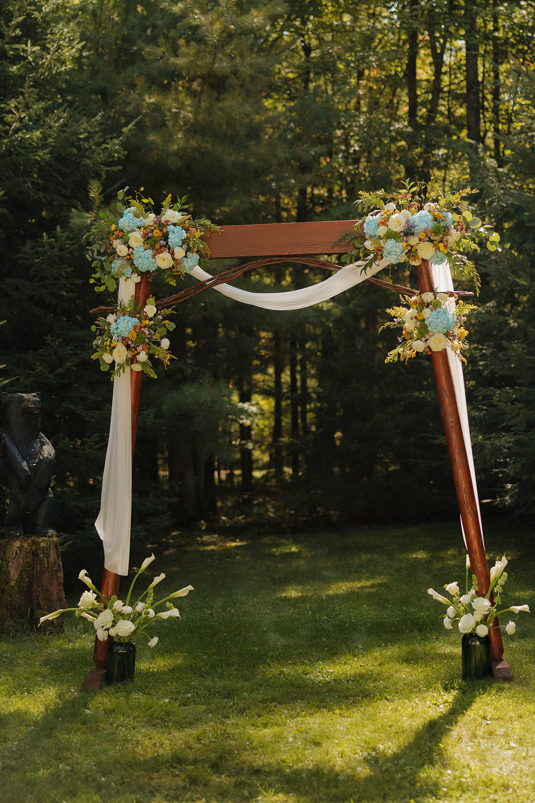 Wooden wedding arch decorated with white fabric and bright floral arrangements for a barn wedding ceremony.
