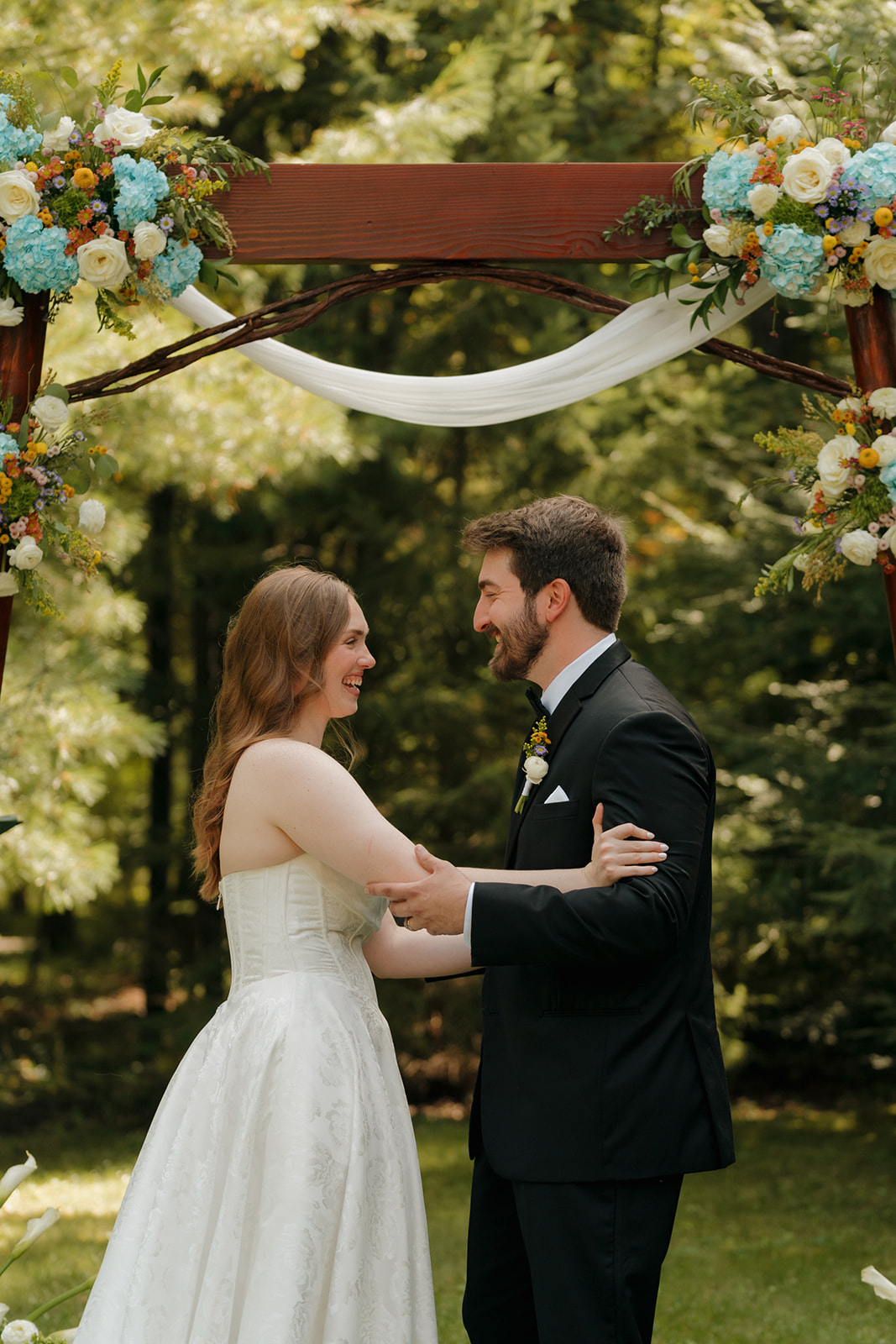 Bride and groom holding hands and laughing under a floral arch in the forest.